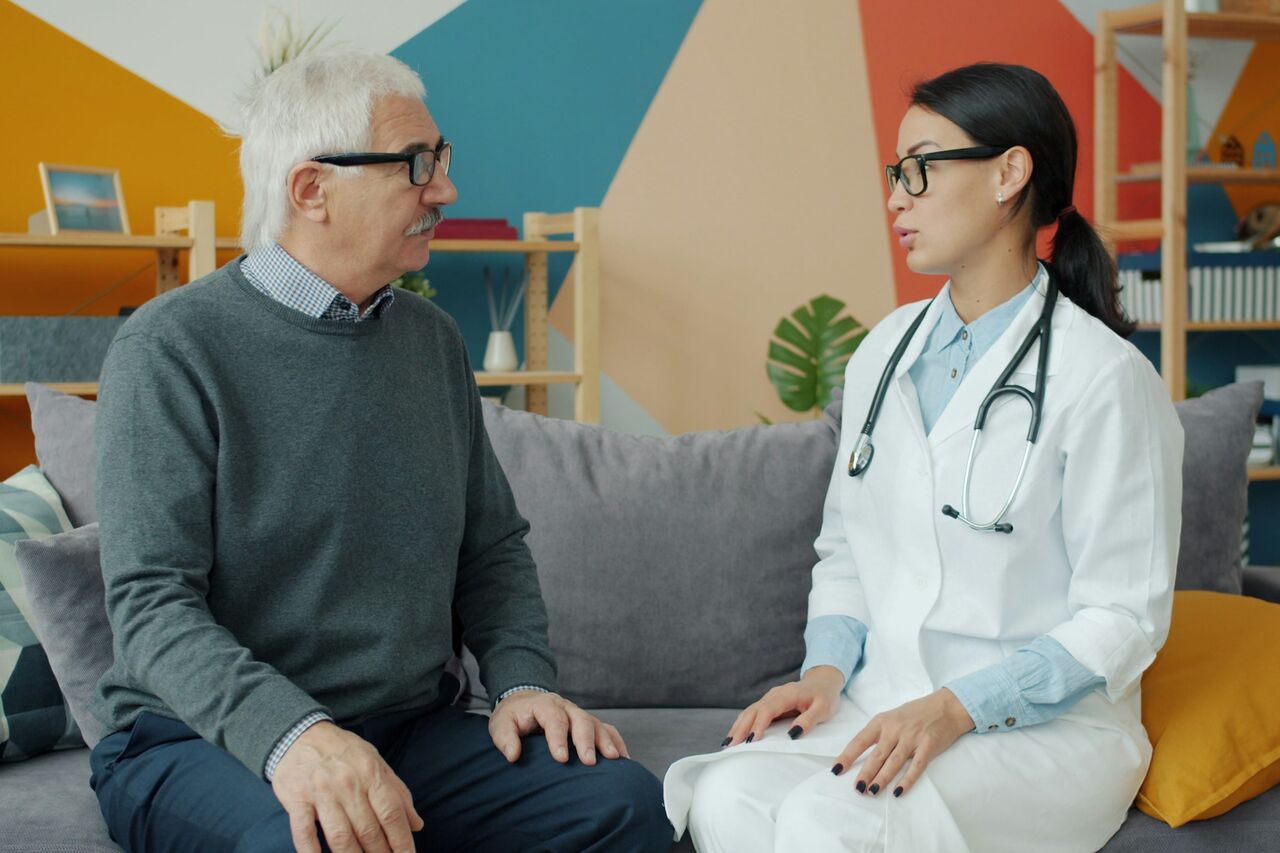 A doctor in a white coat talking with an older patient on a couch
