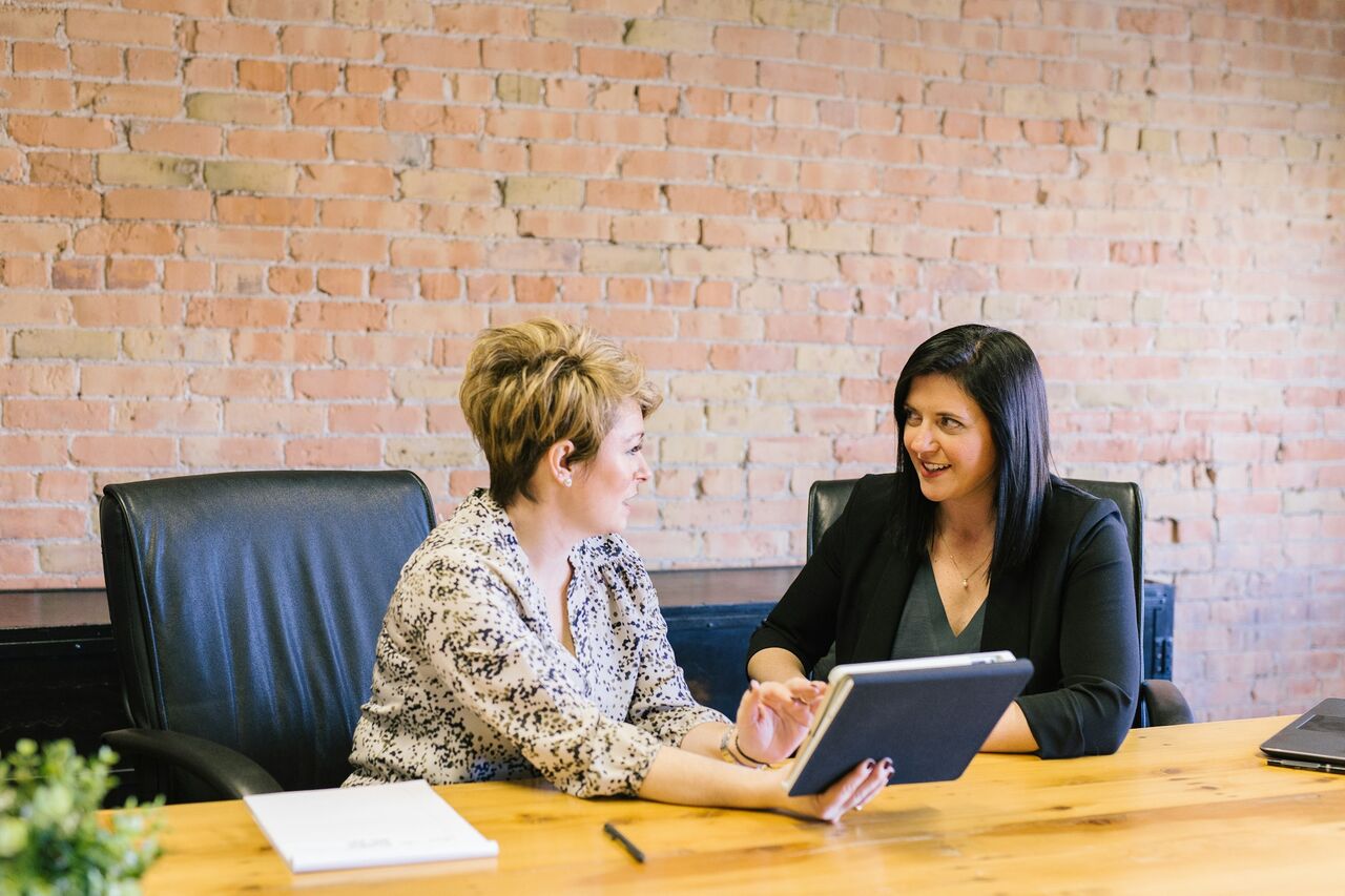Two women at a wooden table looking at a tablet together in a brick-walled office