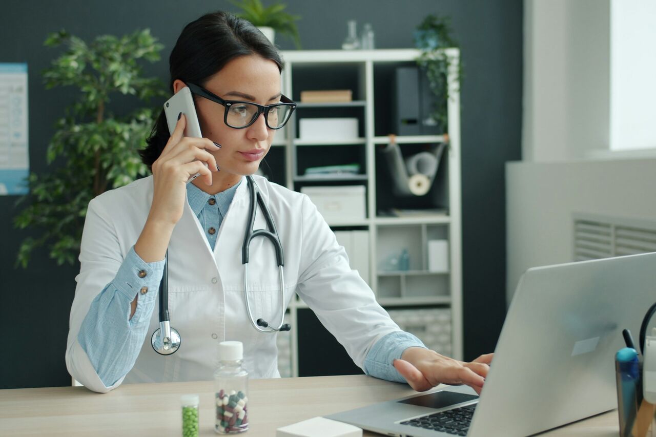 A doctor in a white coat on the phone while typing on a laptop at a desk